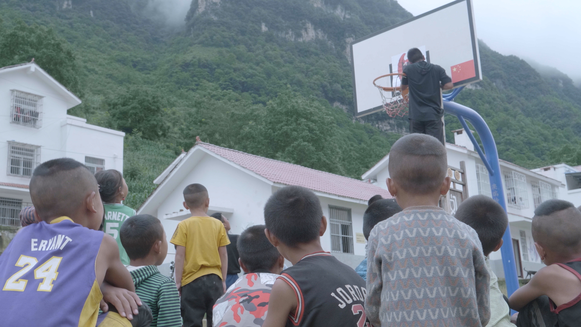 A coach climbs a basketball hoop, a crowd of boys watch him